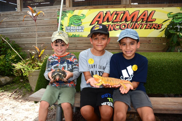 Three children holding reptiles at an animal farm, sitting on a bench with a sign behind them.