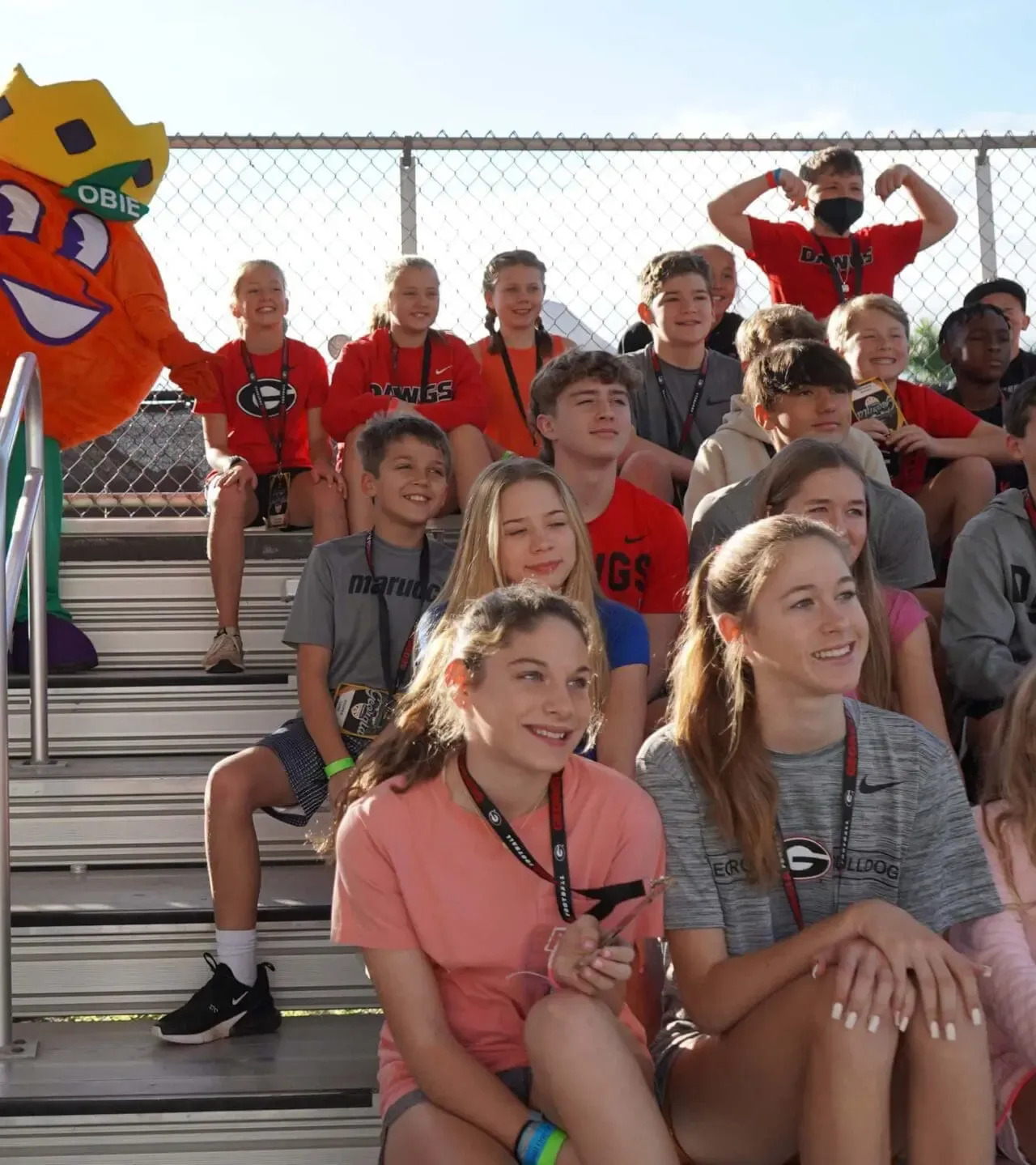 Group of kids sitting in bleachers with a smiling orange mascot nearby.